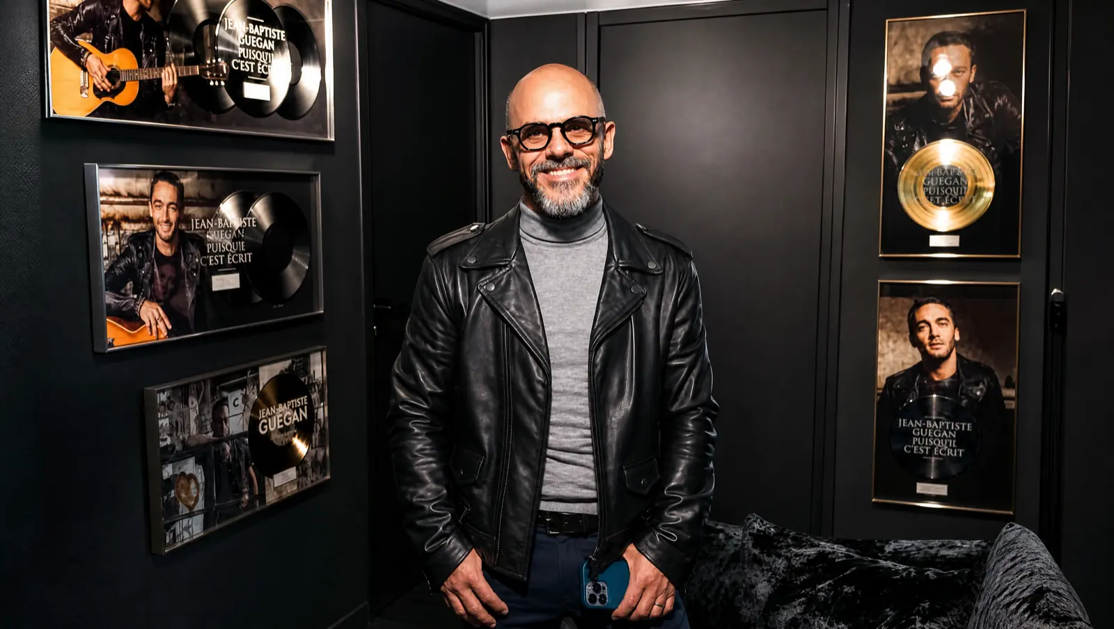 Christophe Porquet in a black leather jacket standing among the framed gold and platinum records for Puisque c’est écrit.